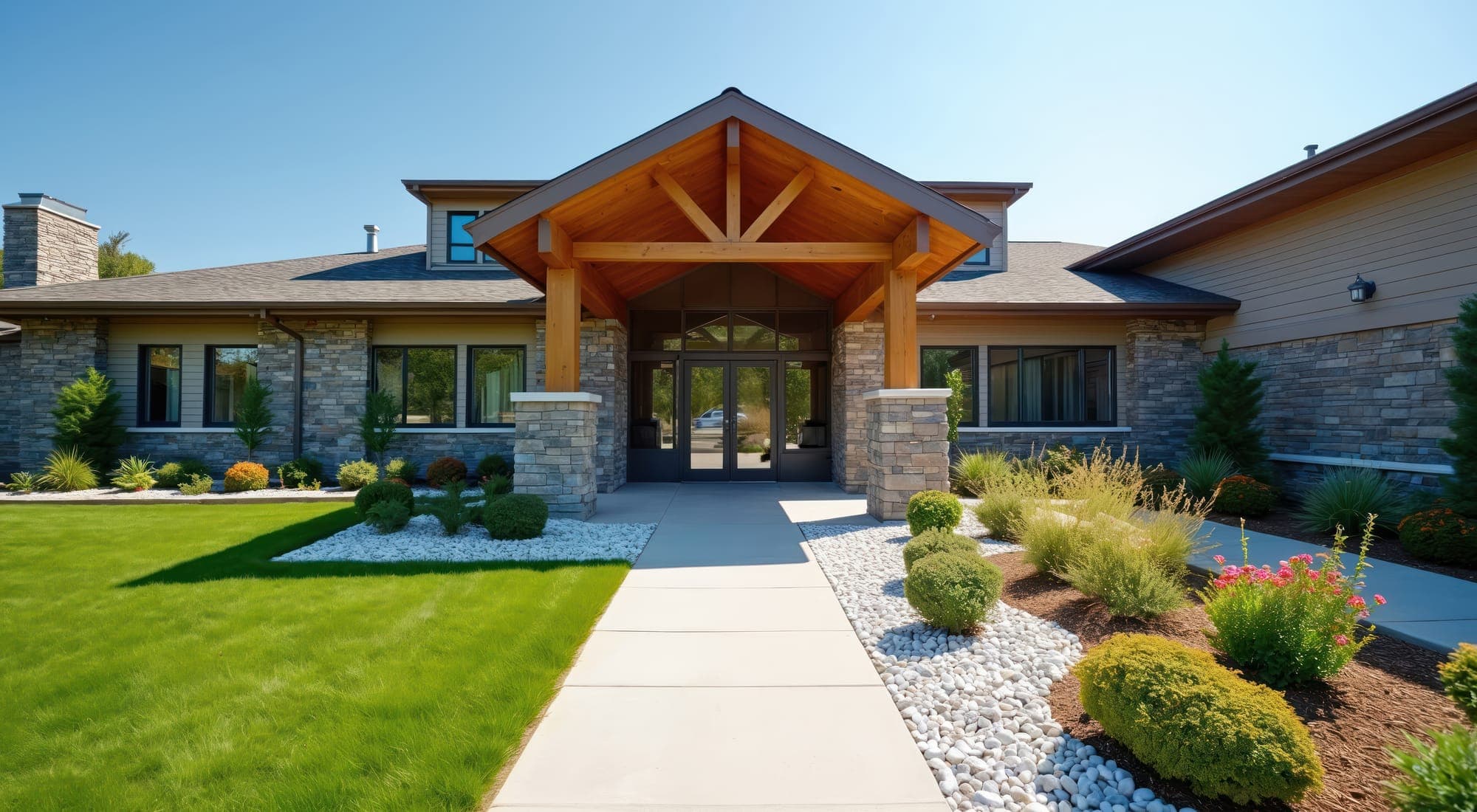 A single-storey care building with a timber-framed entrance canopy, stone piers and a landscaped walk under a clear sky