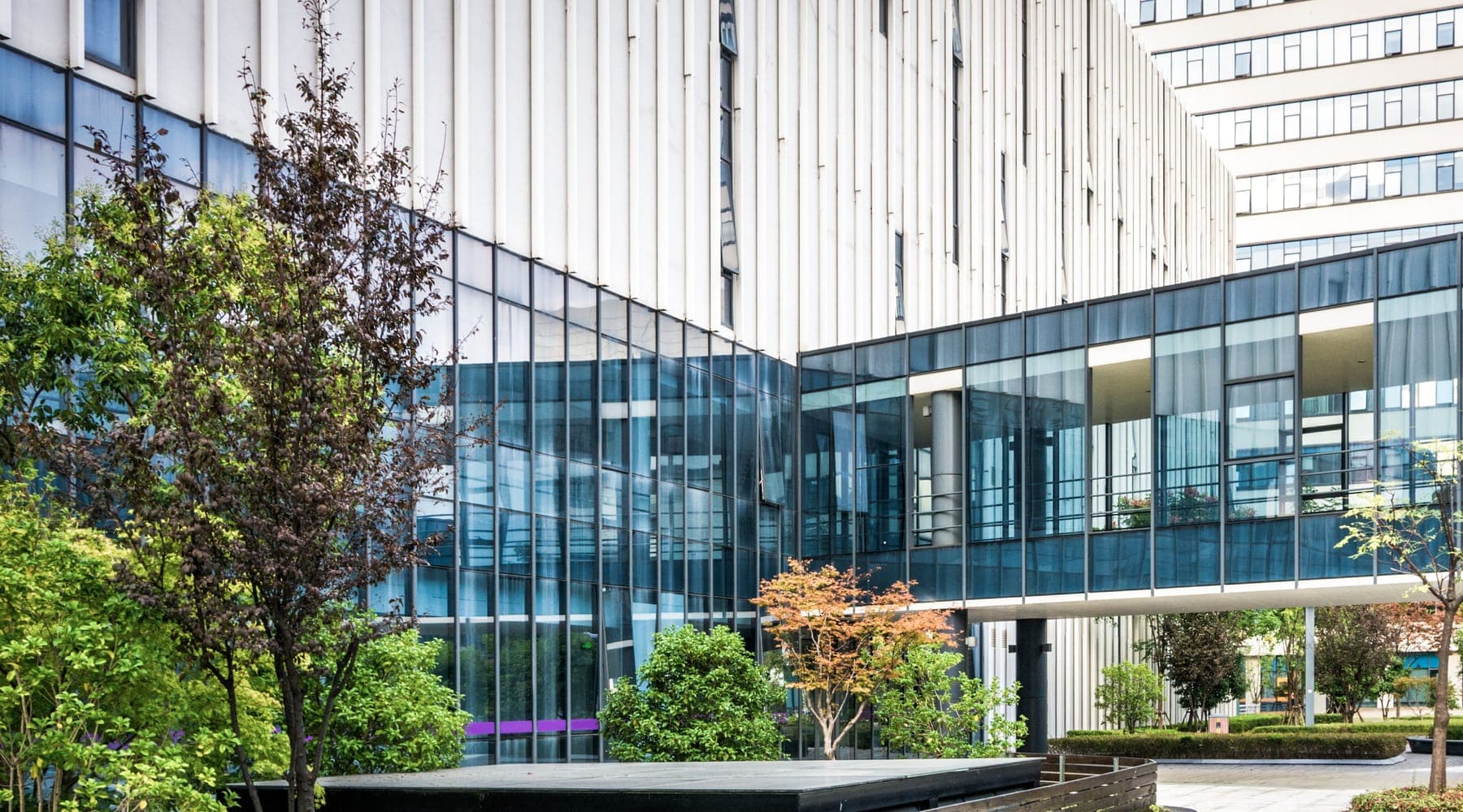 A glass-walled building with an elevated glazed walkway above a landscaped courtyard