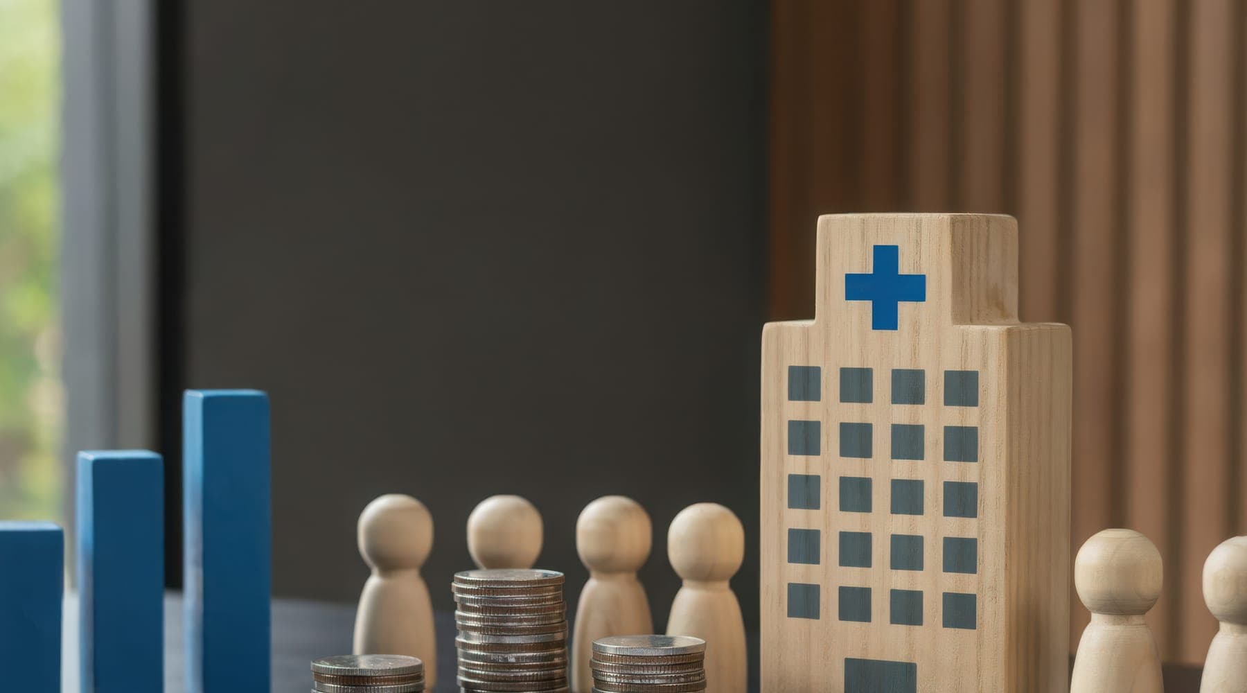 A wooden model of a hospital building beside wooden figures, stacked coins and blue bar-chart blocks on a table