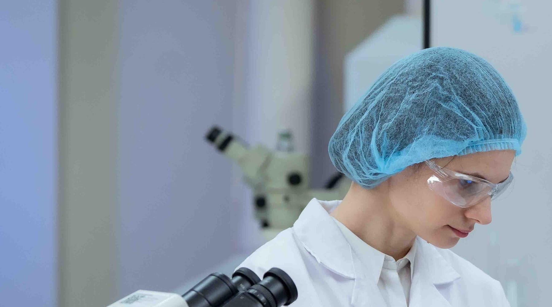 A scientist in a hairnet and safety glasses working at a microscope