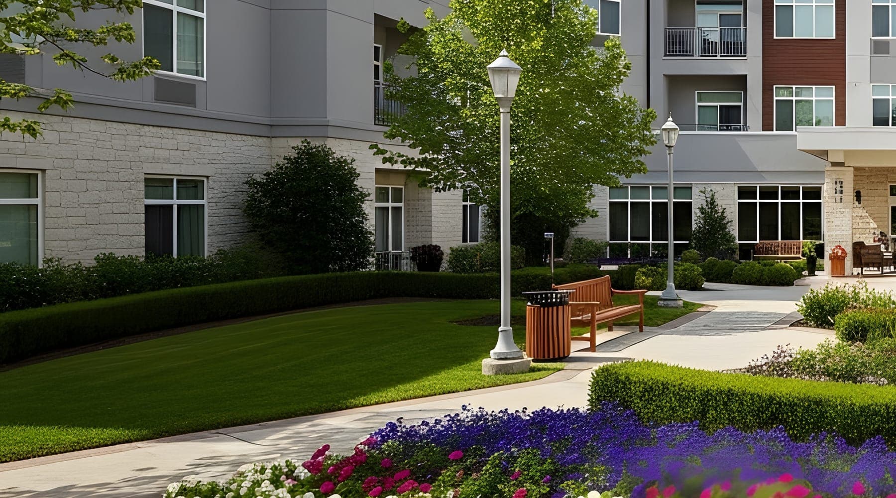 A landscaped courtyard with lawn, benches, lamp posts and flower beds in front of a three-storey residential building