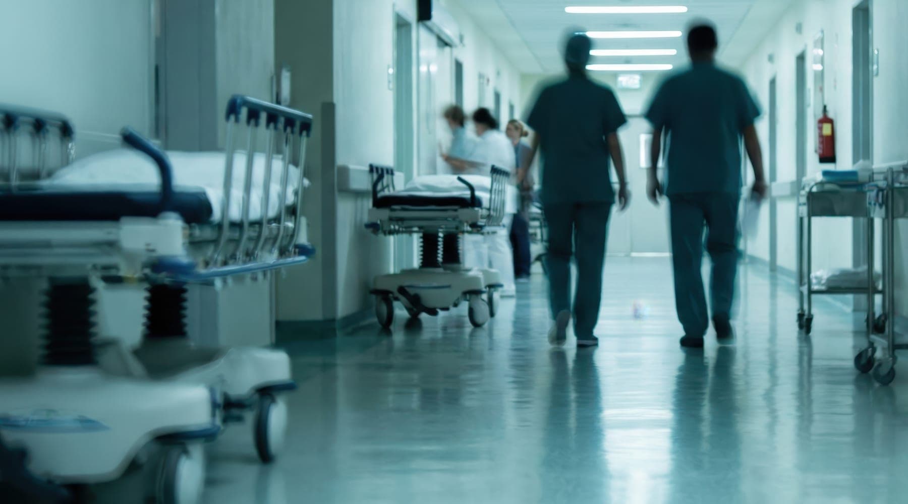 Two clinicians in scrubs walking down a hospital corridor lined with stretchers and equipment carts